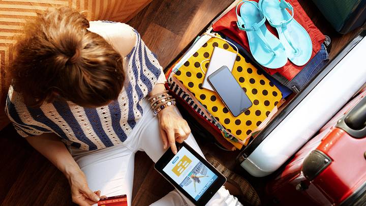 Upper view of young woman in white pants and striped blouse at modern home in sunny summer day book flight on tablet PC near open travel suitcase.