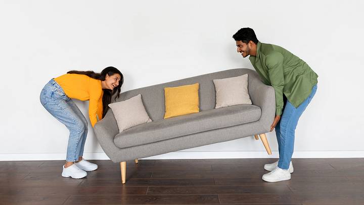 Happy indian spouses moving grey sofa in room over white wall background at their new home, moving to new apartment, full length, free space Happy indian spouses moving grey sofa in room over white wall background at their new home, moving to new apartment, full length, free space
