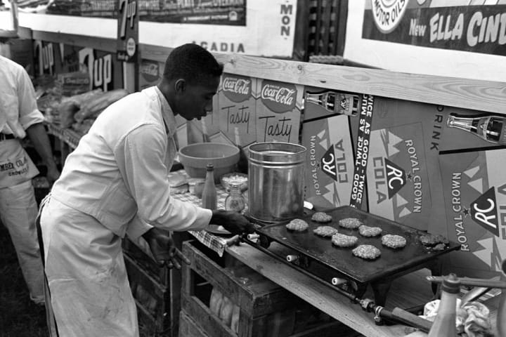 a man in an all-white cook's uniform flips burger patties on a griddle at a concession stand
