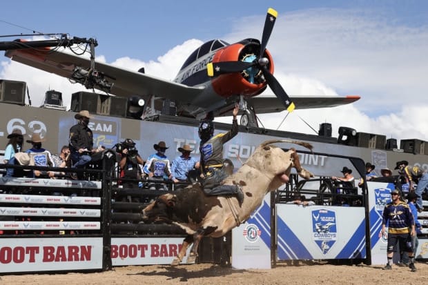 A white and red bucking bull bucking with a cowboy riding him in an arena with an airplane overlooking.