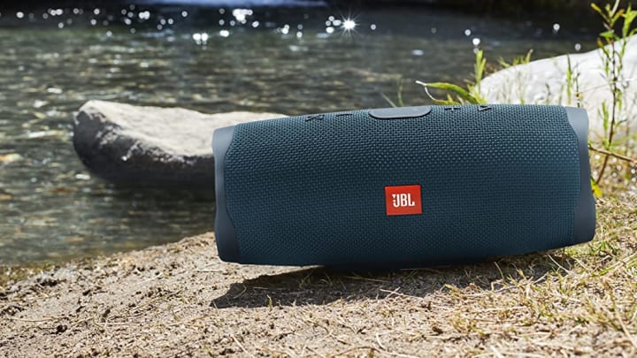 JBL Charge 4 portable Bluetooth speaker on ground by water.