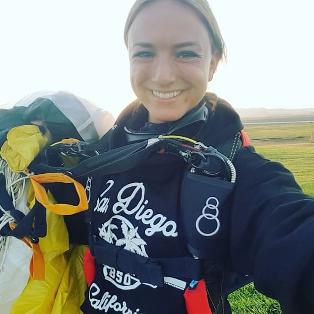 A female skydiver in black skydiving clothing smiling at the camera as she holds a yellow and white parachute on the grass.
