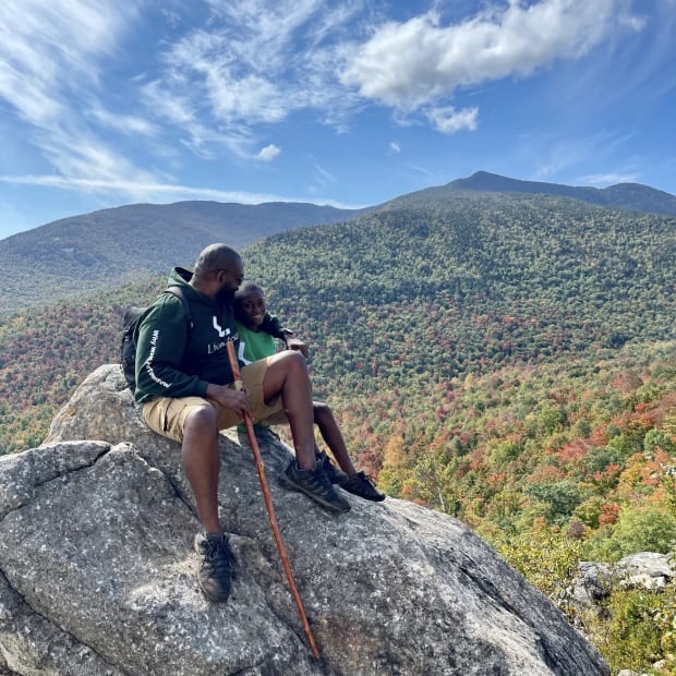 Clifton Harcum with his arm around his son on a rocky summit with a beautiful autumn background in the Adirondack mountains.