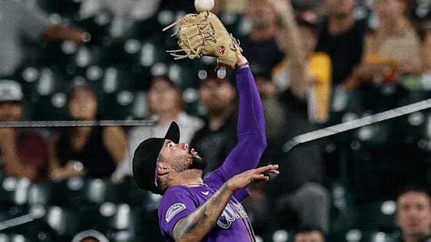 A baseball player in a purple jersey and black hat reaching up into the air with his brown glove to catch a white baseball.