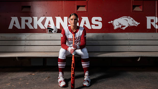 A softball player in a red and white jersey sits on a bench holding a bat