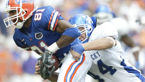 A football player wearing a white jersey and blue helmet tackles a player wearing a blue jersey and orange helmet.