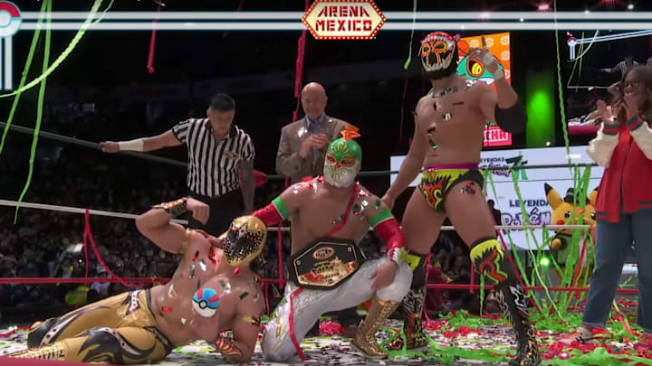 Three Luchadors posing in the ring with a championship as streamers fall from above.