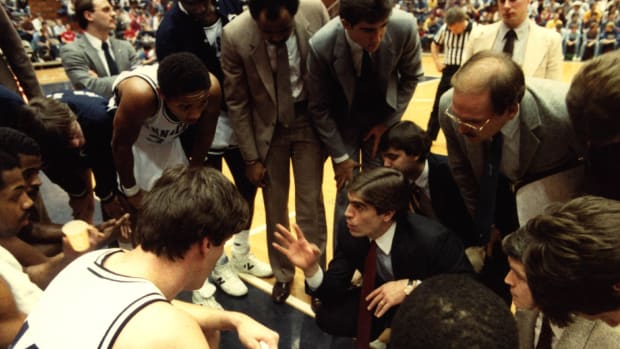 Former Penn State men's basketball coach holds up his hand as he speaks to players in a huddle around him.