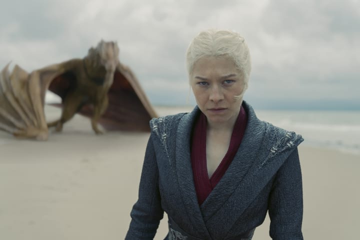 Rhaenyra Targaryen glares at something offscreen, while her dragon Syrax looms behind her. They're on a beach.