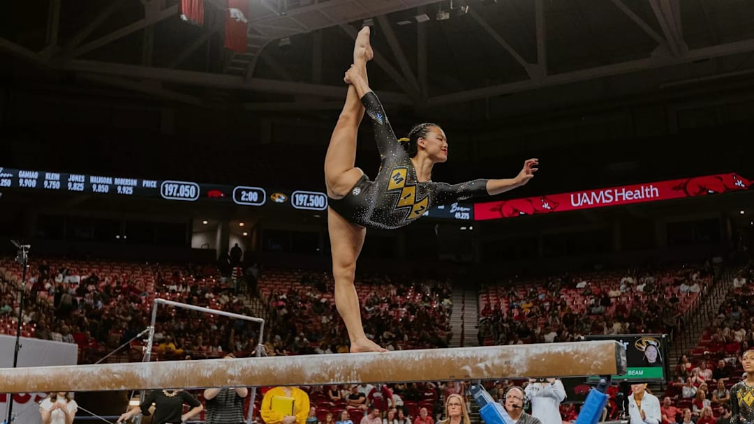 United States; Missouri gymnast Helen Hu competes on beam, holding her leg up behind her while displaying a Tiger claw with her hand.