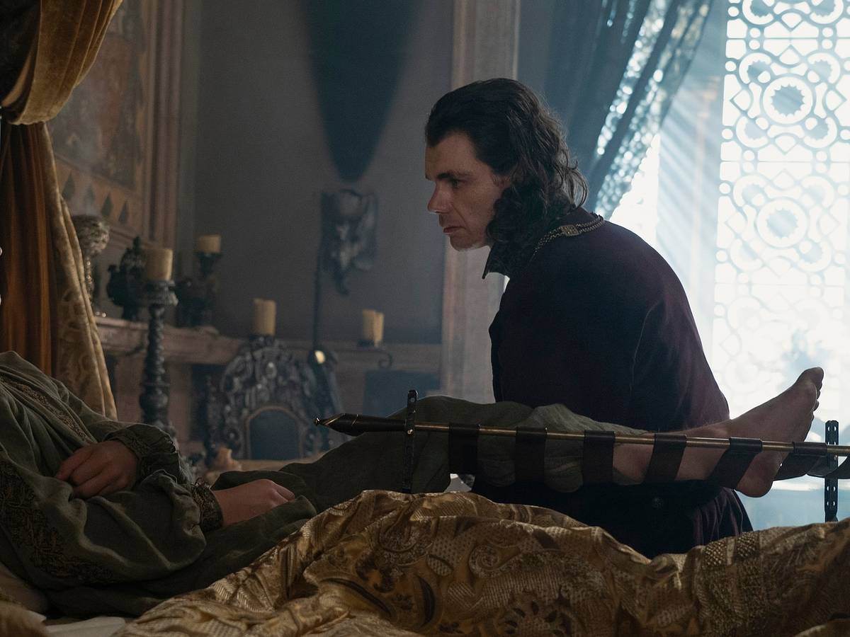 A man with shoulder-length blonde hair (Aegon) sits up in bed wearing a green tunic. His leg is propped up and he has a golden blanket. A man with shoulder-length brown hair (Larys) sits by his bed, framed to his right. Behind them are various decorations like a painting, candles, and an ornate chair.