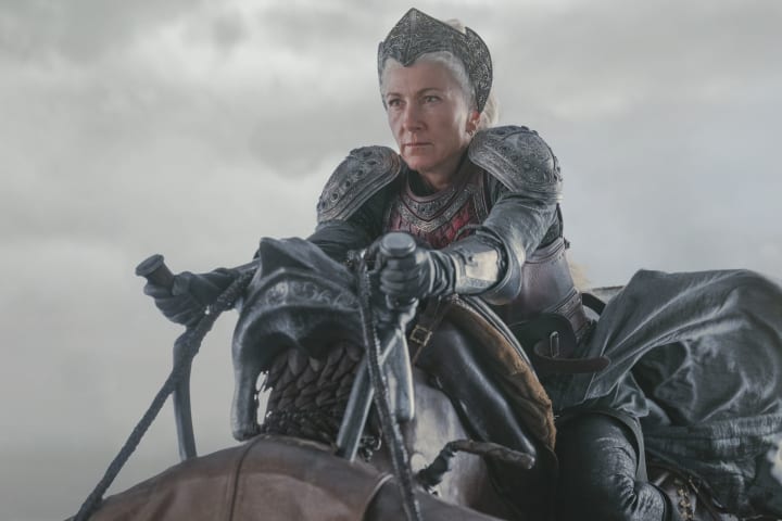 Rhaenys Targaryen (Eve Best) sits in her dragon saddle, wearing red and black armor and an ornate battle crown.