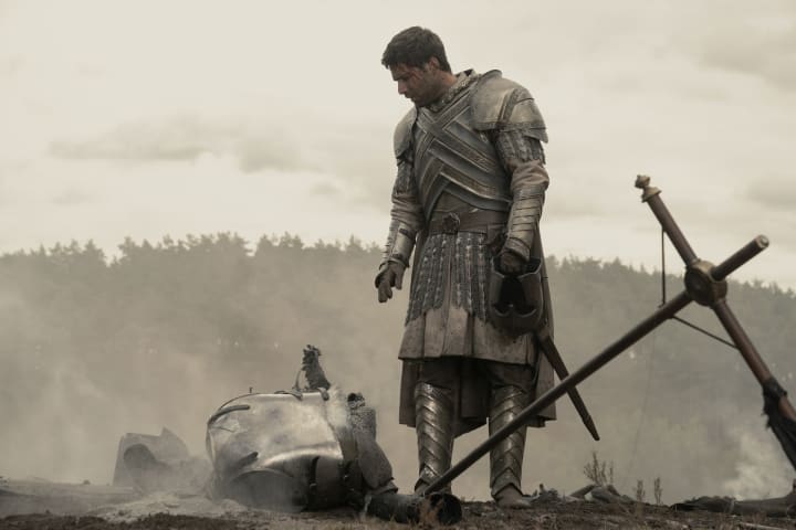 Ser Criston Cole (Fabien Frankel) stands on a charred battlefield in his Kingsguard armor, looking at an armored corpse.