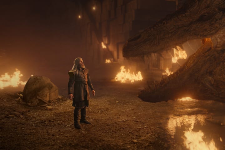 Hugh stands before the dragon Vermithor, while fires burn around them.