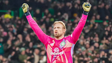 Celtic goalkeeper Kasper Schmeichel stands on the pitch with both hands raised in fists, wearing a pink jersey with yellow gloves. 