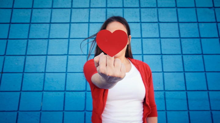 woman stands in front of a blue wall and flips off the camera while concealing it with a paper heart