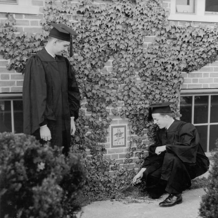 two cap-and-gown-clad senior boys planting ivy below an ivy-surrounded plaque inscribed with "B" and "1943"