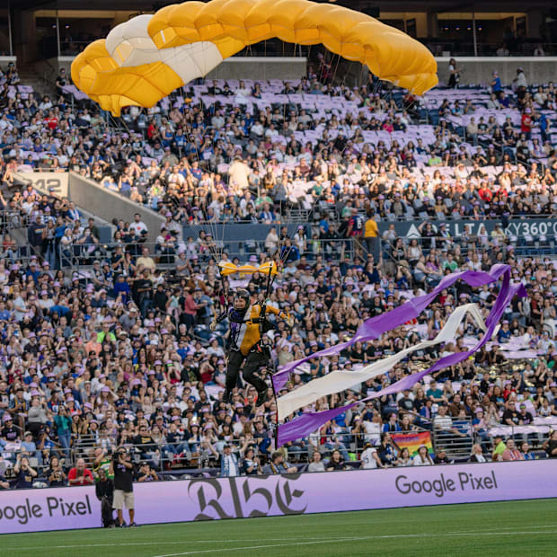 Skydiver with a yellow and white parachute landing in Lumen Field with an excited crowd in the background.