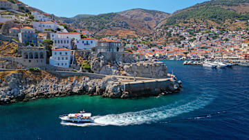 Drone footage image of a boat in front of houses on cliffs in Hydra, Greece
