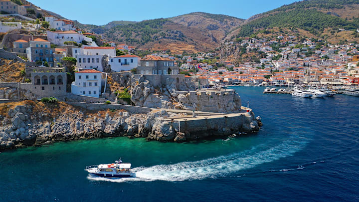 Drone footage image of a boat in front of houses on cliffs in Hydra, Greece