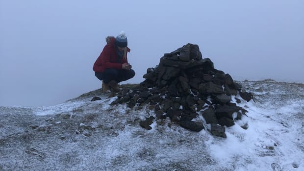 A woman in red sits next to brown rocks atop a mountain peak