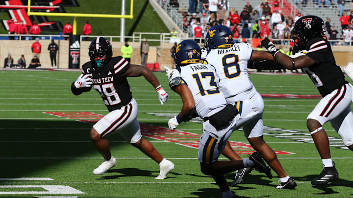 Nov 30, 2024; Lubbock, Texas, USA;  Texas Tech Red Raiders running back Tahj Brooks (28) runs the ball against West Virginia Mountaineers defensive back Dontez Fagan (13) and defensive back Tyrin Bradley Jr. (8) in the second half at Jones AT&T Stadium and Cody Campbell Field. Mandatory Credit: Michael C. Johnson-Imagn Images