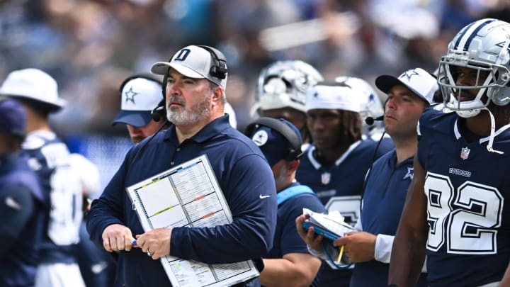 A football coach stands on the sidelines, holding a playbook, surrounded by team members in uniforms.