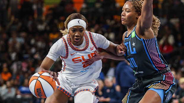 A basketball player in a white uniform that says "Sun," dribbling a white and orange basketball past another player.