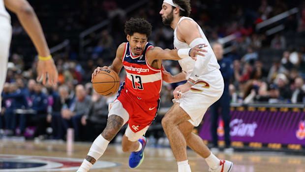 A basketball player wearing a red uniform dribbling a basketball next to another player wearing a white uniform.