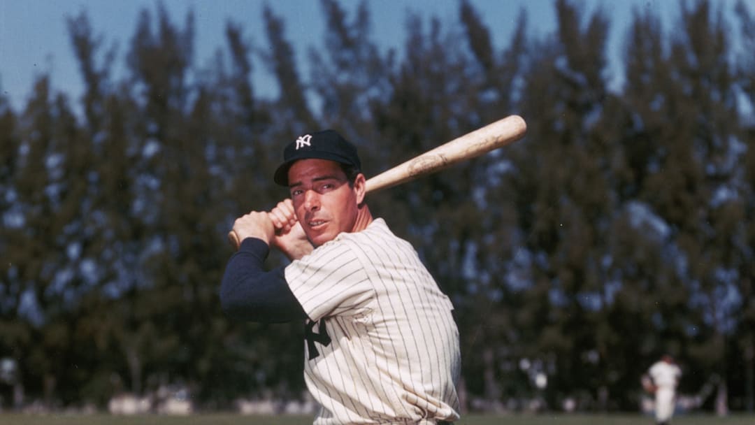 Slugger Joe DiMaggio poses for a picture in an empty field. Standing upright, he holds the bat in the ready position. His jersey billows off his large torso.