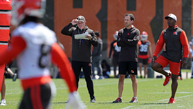 Two men in brown jackets and black pants standing on a green field next to players in brown and red uniforms.