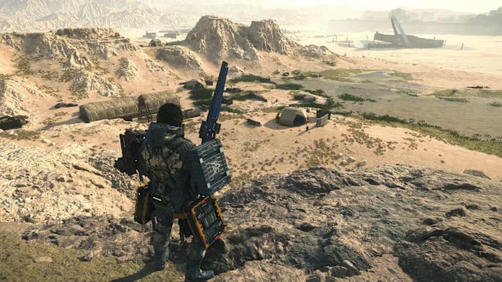 Sam stands looking down over a desert base, sniper rifle ready, in Death Stranding 2. 