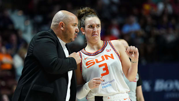 A female basketball player in a white uniform saying "Sun 3" in orange, standing next to a man in a black suit.