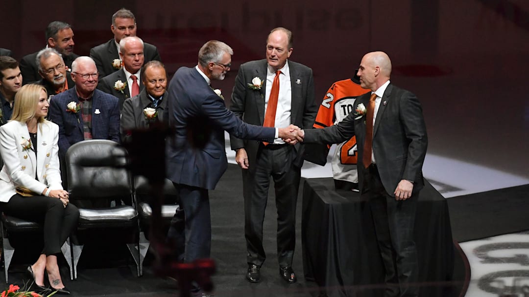 Nov 16, 2021; Philadelphia, Pennsylvania, USA; Former Philadelphia Flyers Paul Holmgren, left, shakes hands with Rick Tocchet after they were inducted to the Flyers Hall of Fame during a ceremony before game against the Calgary Flames at Wells Fargo Center. Dave Scott, Comcast Spectacor Chairman and CEO, and Governor of the Flyers stands between them.