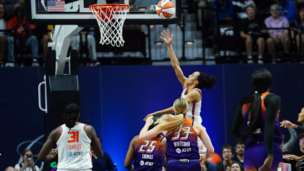 A female basketball player in a white uniform shooting an orange and white basketball into the air toward the basketball hoop
