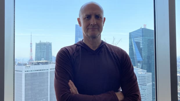 Tom Hovasse, coach of the Japan men's national basketball team, stands in front of window with the Tokyo skyline behind him.