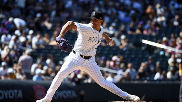 A baseball player in a white uniform that says "Rockies" while wearing a black hat, pitching a white baseball during a game