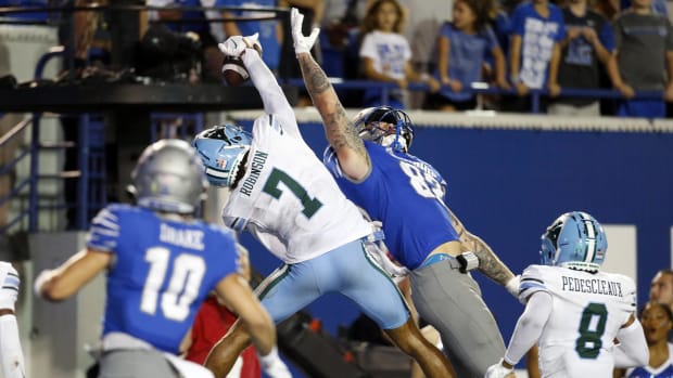 A defender breaks up a pass in a blue helmet and pants and white jersey with green numbers over a player in blue jersey