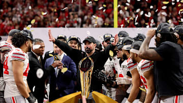 Ryan Day stands with his Ohio State Buckeyes football team behind the 2025 CFP national championship trophy after beating Notre Dame, 34-23.