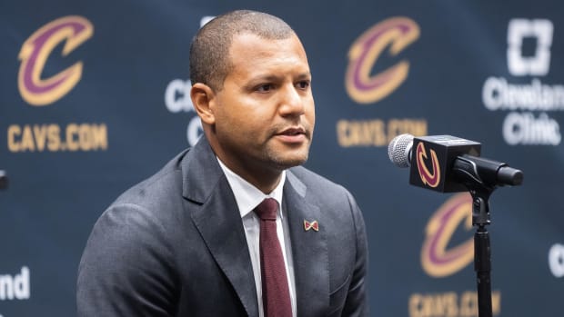 A basketball executive wearing.a black suit and maroon tie while sitting in front of a microphone.