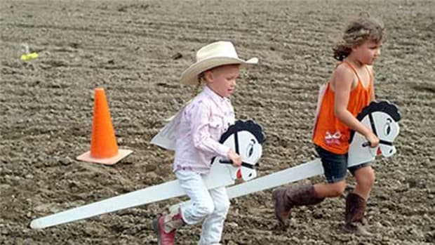 Two kids on stick horses racing in the arena dirt.