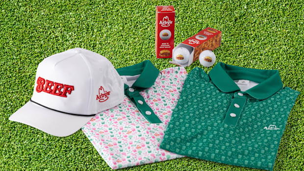 College golf NIL collection with white hat, pack of golf balls, and green and pink polos on golf turf.