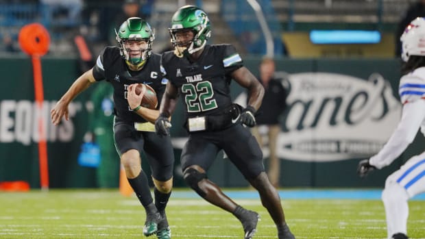 Two football players in black jerseys with green numbers and black helmets run down the field with the football.