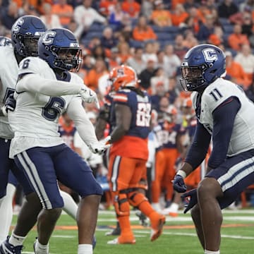 Two University of Connecticut football players celebrating on the field, wearing white uniforms with blue accents, amidst an excited crowd in the background.