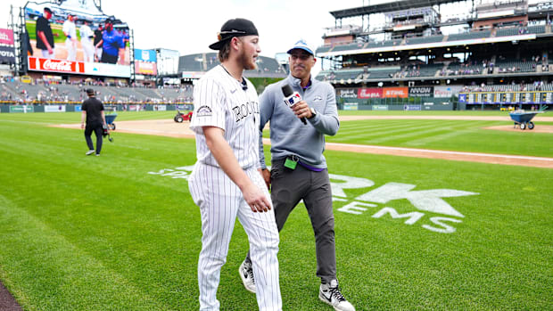 Colorado Rockies player talks into a mic wearing a white and black pinstripe jersey and black backwards hat.