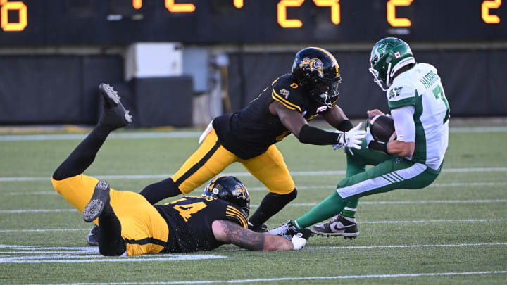 Jun 16, 2024; Hamilton, Ontario, CAN; Saskatchewan Rough Riders quarterback Trevor Harris is tackled by Hamilton Tiger Cats linebacker Ray Wilborn (0) and defensive tackle Casey Sayles (44) in the second quarter at Tim Hortons Field. Mandatory Credit: Gerry Angus-USA TODAY Sports