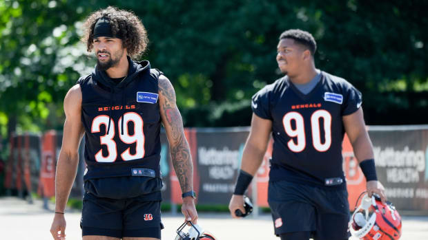 Two football players walk to practice wearing black jerseys with white numbers and orange outline and black pants.