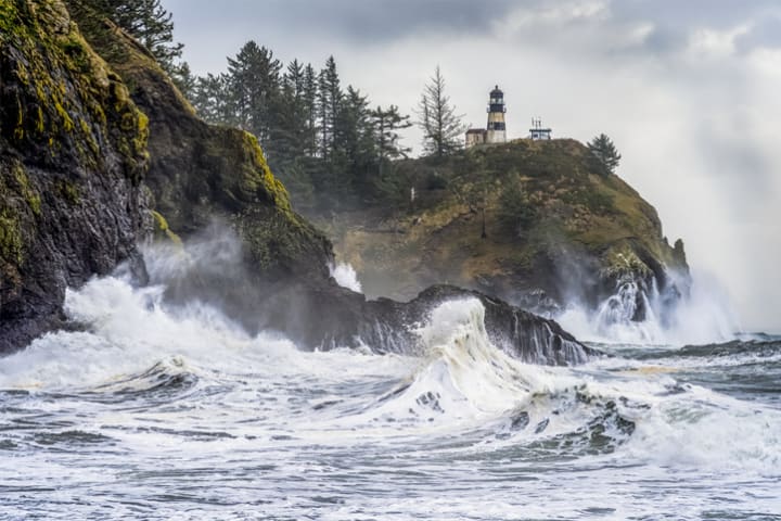 Waves crashing against the coast in the Pacific Northwest with a lighthouse on a cliff.