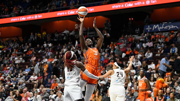 A basketball player in an orange uniform jumping up to shoot a basketball while two players in white uniforms guard her