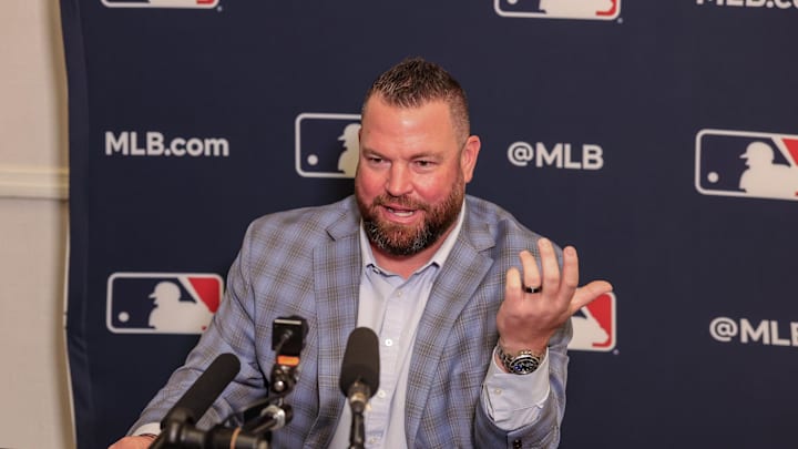 A man sitting down in a light blue checkered suit, speaking in front of a microphone while in front of an MLB backdrop.